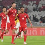 Empat pemain sepak bola berbaju merah berada di lapangan, salah satu dari mereka mengacungkan simbol damai. Latar belakang menunjukkan stadion dengan penonton.